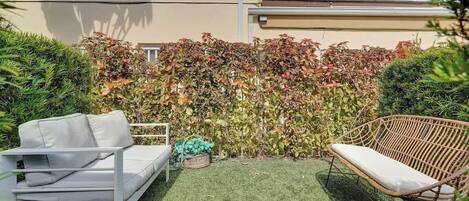 Un patio con un sofá blanco y una silla de mimbre, rodeado por un seto con follaje otoñal.