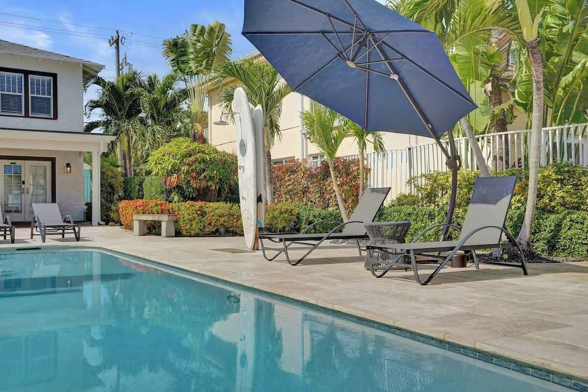 Una piscina con lettini e un ombrellone grande, circondata da piante tropicali e un recinto bianco.