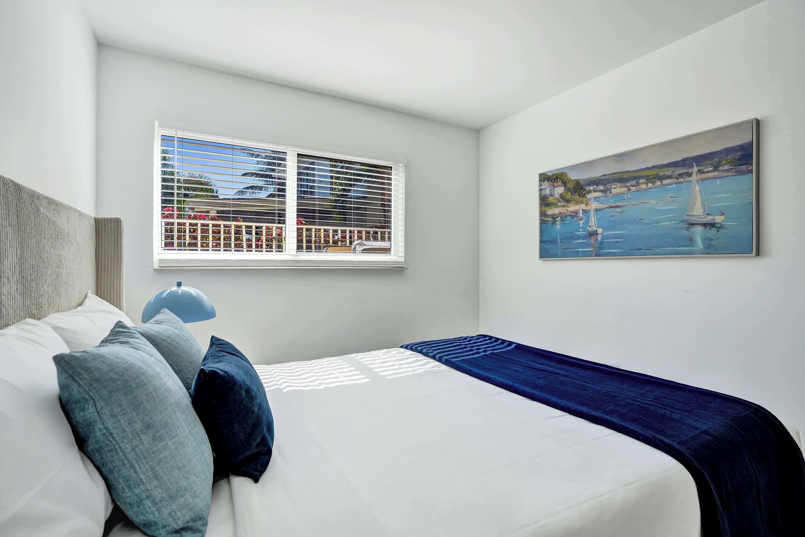 A bedroom with a bed, pillows, a window with blinds, a painting of a harbor, and a lamp.