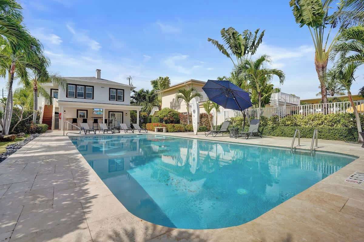A swimming pool with lounge chairs and umbrellas, surrounded by palm trees and a building with a white facade.