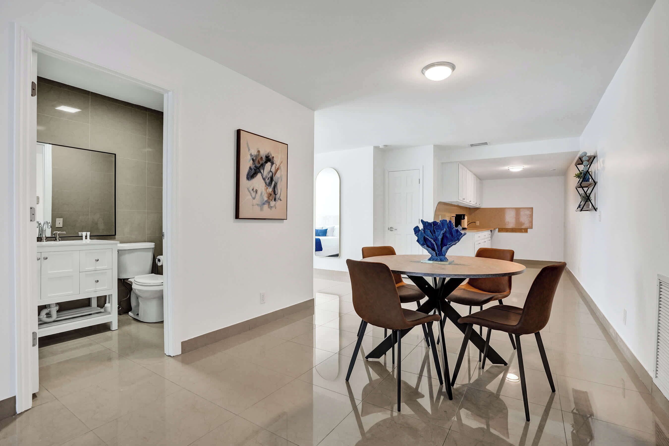 A modern kitchen with white cabinets, a dining area with a round table and chairs, and a visible bathroom with a toilet and sink.