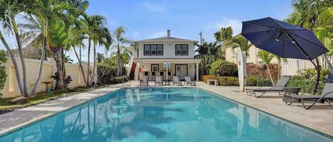 A swimming pool with lounge chairs and an umbrella, surrounded by palm trees and a two-story house.