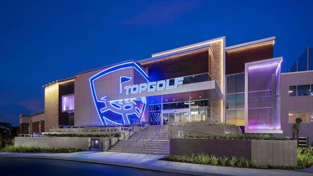 En modern byggnad med en tydlig skylt som visar texten ”TOPGOLF”, en trappa som leder till entrén och en väl upplyst fasad.