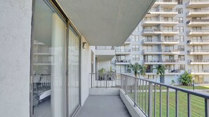 En balkong med en glasdörr, ett metallräcke och utsikt över en hög byggnad med flera våningar och balkonger.