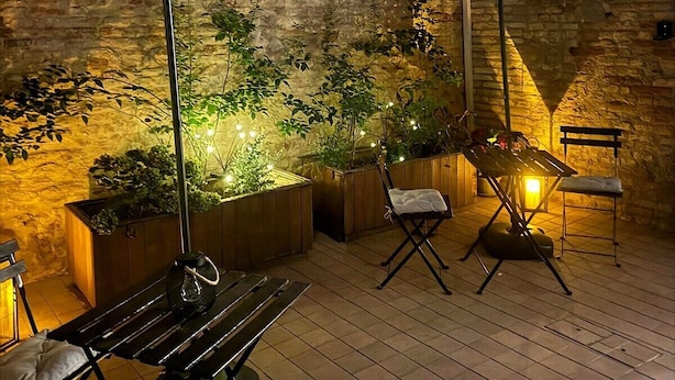 Une terrasse extérieure avec des tables et des chaises en bois, des parasols et des plantes en pot, le tout adossé à un mur de pierre.