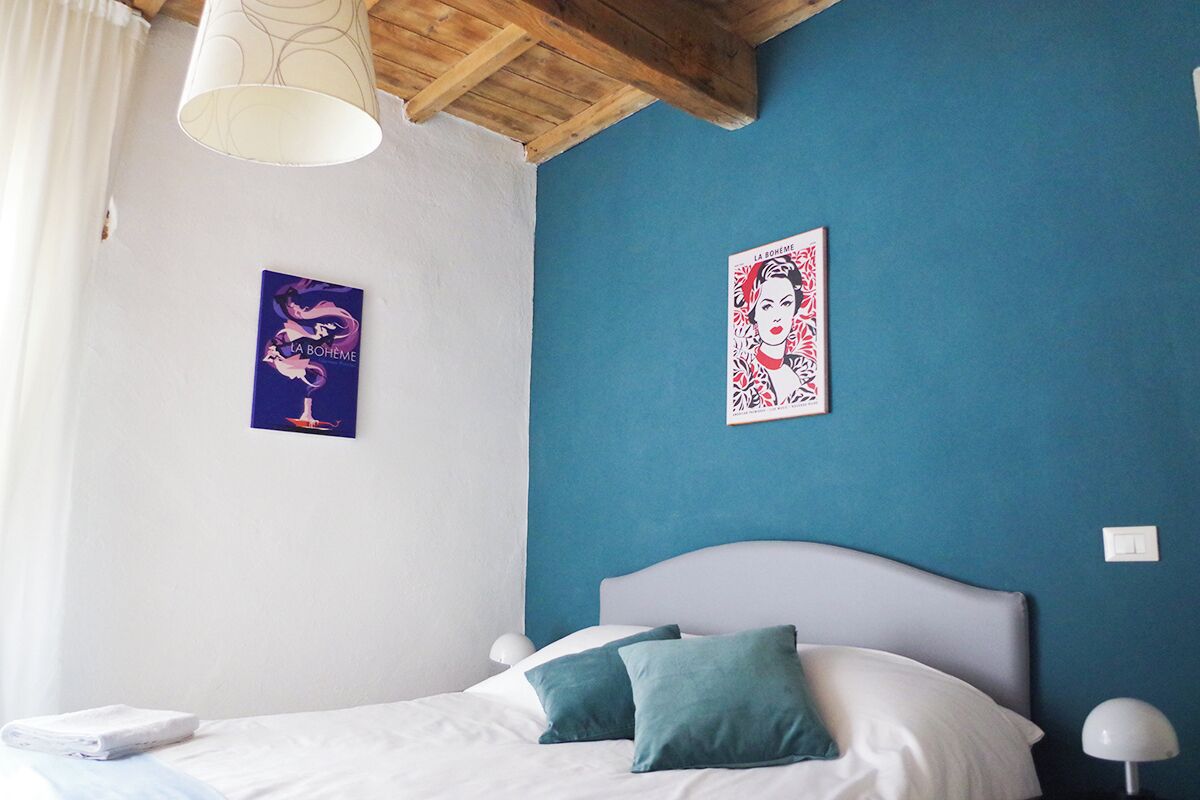 Un dormitorio con cama, almohadas, una mesita de noche, lámpara y dos cuadros en la pared.