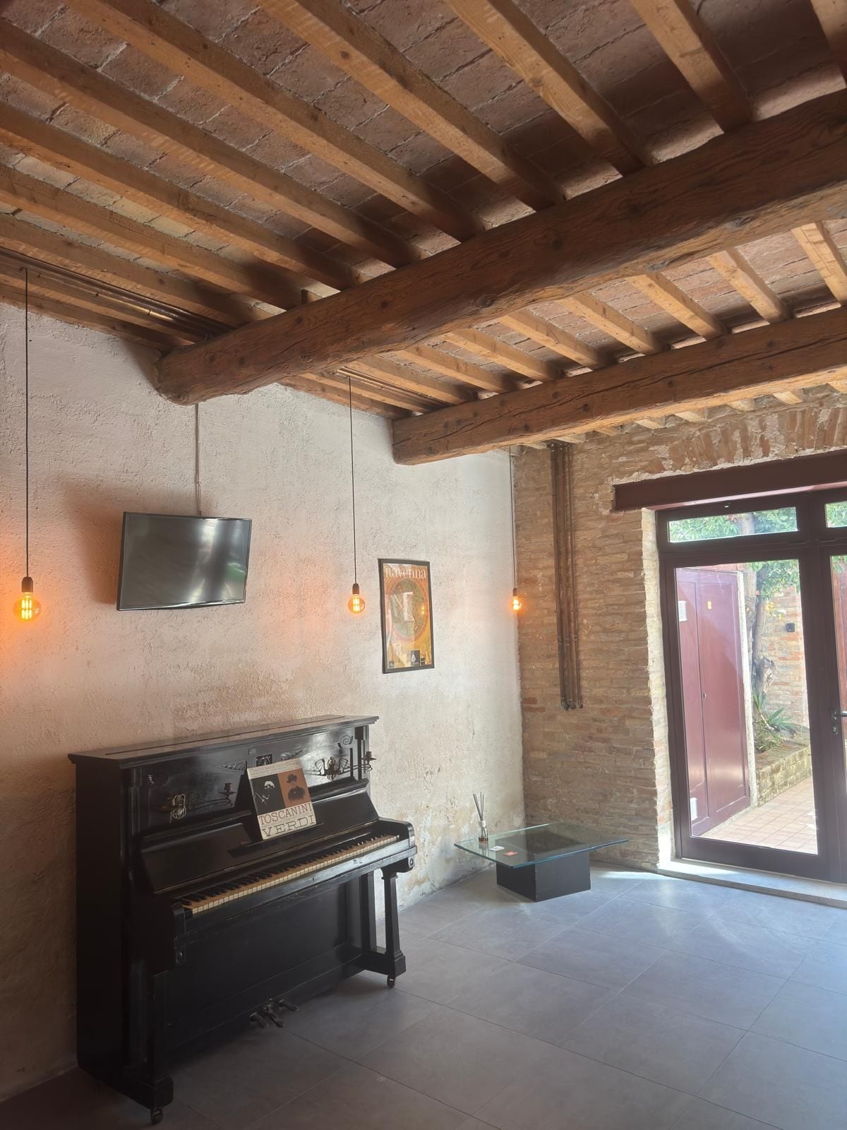 Una habitación con vigas de madera a la vista, un piano, un televisor de pantalla plana y una puerta que da al exterior.