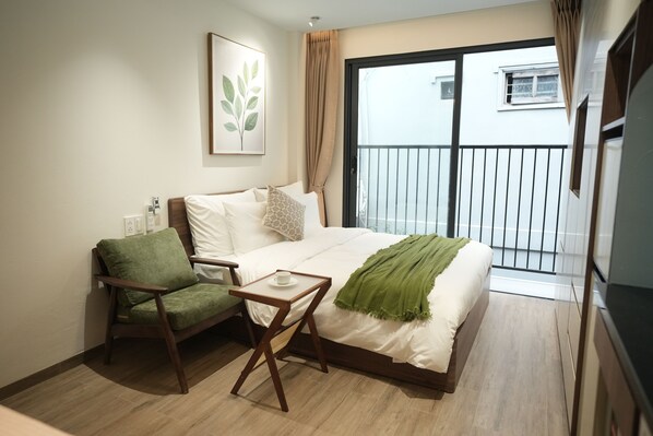 Une chambre d’hôtel avec un lit, un fauteuil vert, une petite table, un tableau encadré au mur et un balcon avec une balustrade.