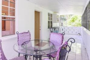 A covered patio area with a glass table and chairs, a door leading to an interior space, and a view of greenery outside.