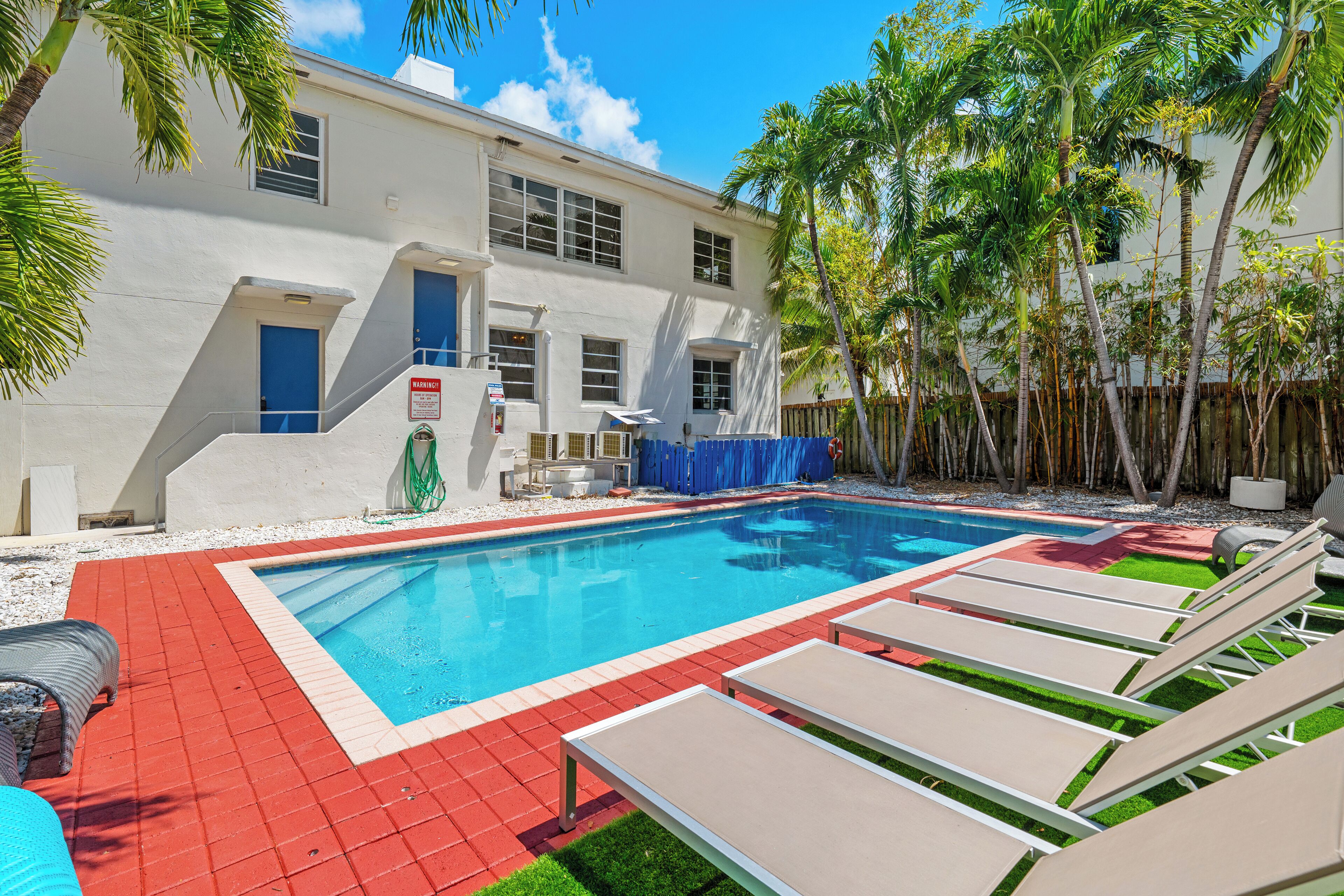A swimming pool with lounge chairs, a white building with blue doors, and palm trees.