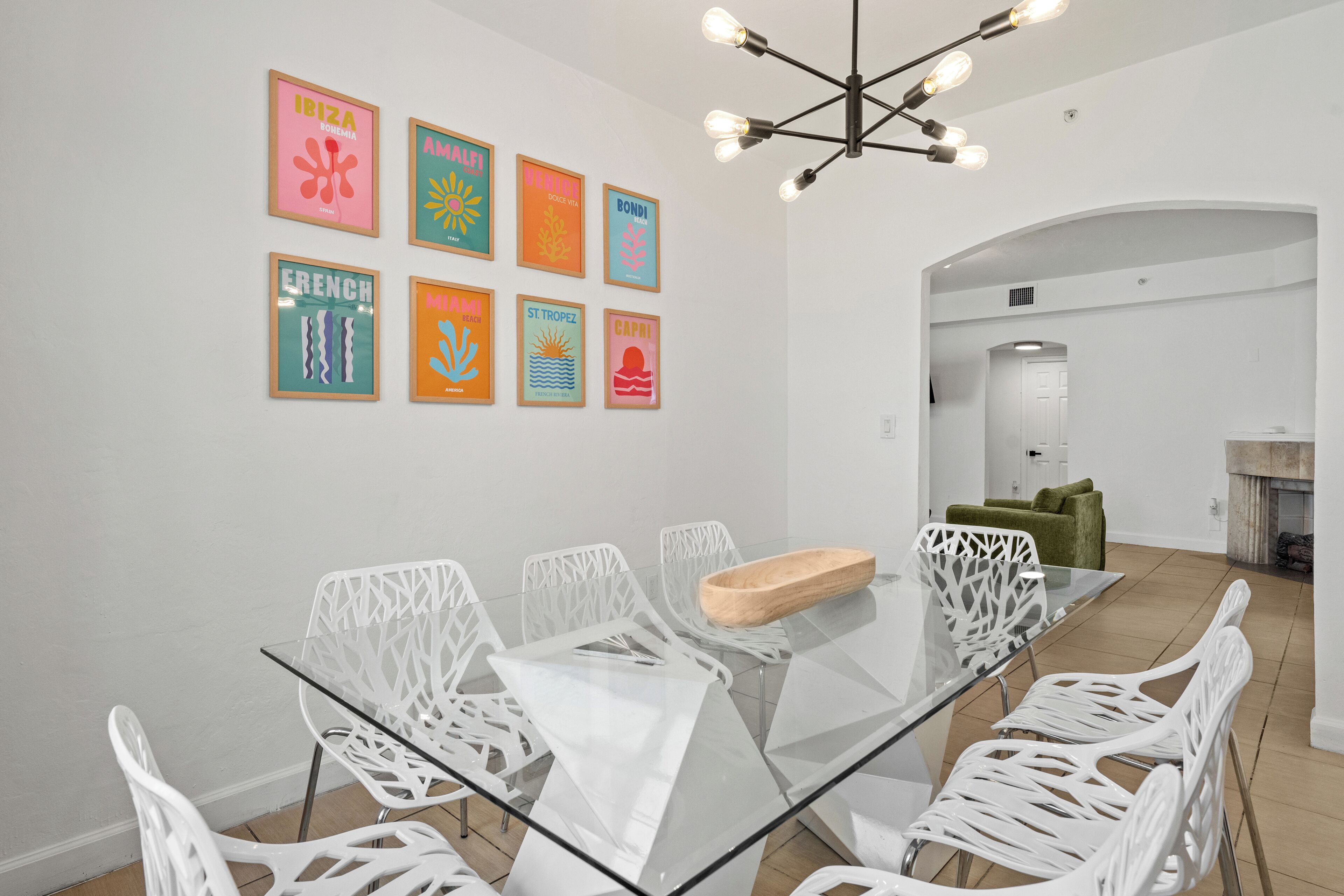 A modern dining area with a glass table and white chairs, surrounded by a white wall with colorful artwork.
