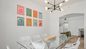 Un espace repas moderne avec une table en verre et des chaises blanches, entouré d’un mur blanc orné d’œuvres d’art colorées.