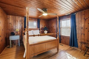 Ein Bett mit Baldachin in einem Zimmer aus Holz, mit Deckenventilator, Nachttisch mit Lampe und einem kleinen Tisch mit Stuhl.