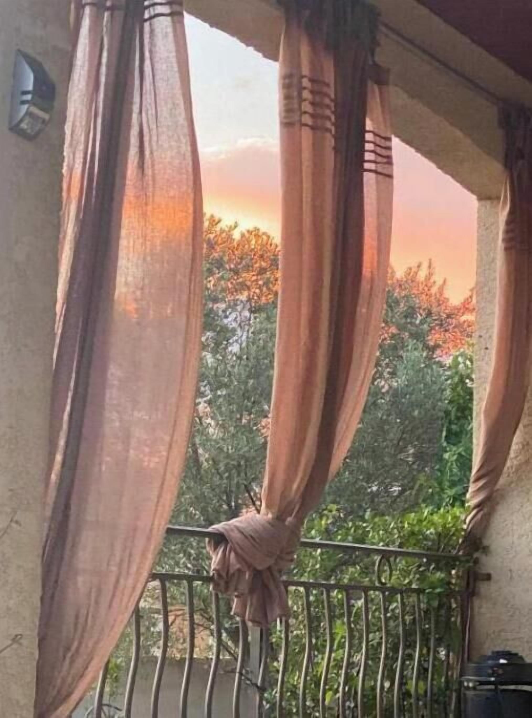 A balcony with sheer curtains, a metal railing, and a view of trees and a sunset.