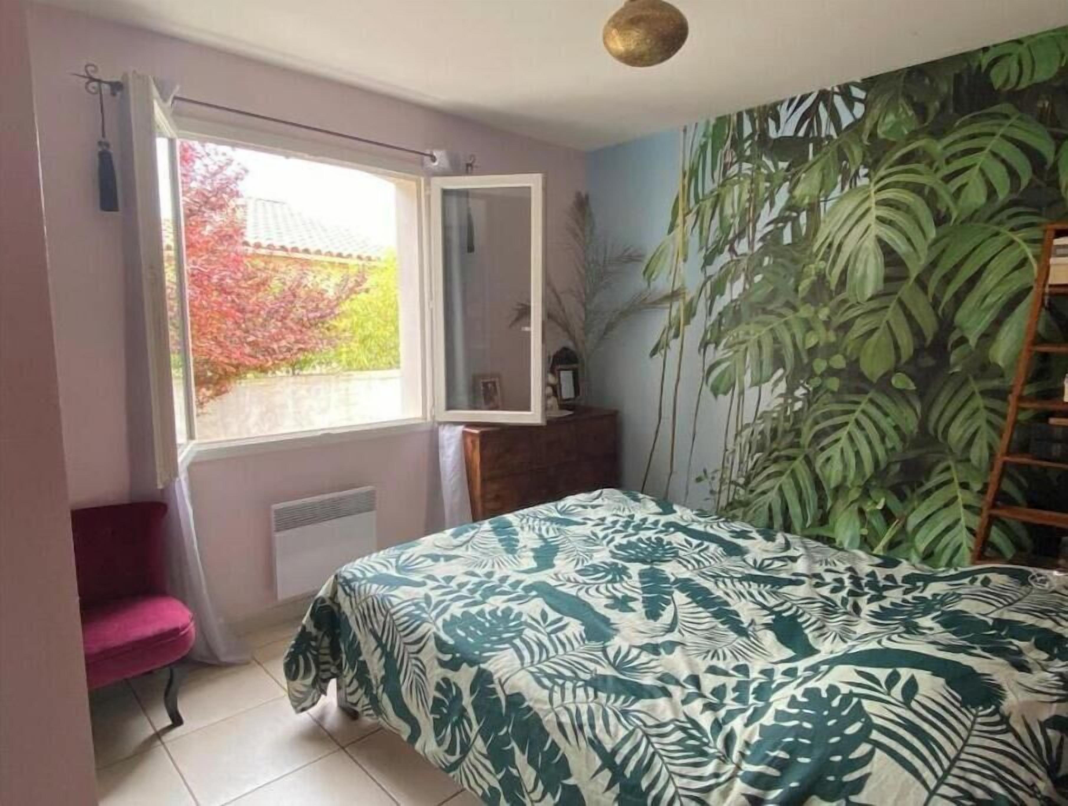Une chambre à coucher avec un lit, une fenêtre, une chaise, une commode et un mur orné d’un motif végétal.