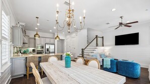 A modern kitchen with white cabinets, a dining area with a wooden table, and a living room with blue sofas. - Community Pool and Hot Tub Access | LSV (Santa Rosa Beach)