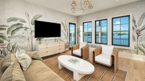 Una sala de estar con televisor de pantalla plana, muebles de mimbre, un tapete estampado y ventanas amplias con vistas.