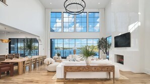 A spacious living room with a large dining table, comfortable seating, a fireplace, and large windows offering a view of the outdoors. - Newly Built | Pool | LSV | YOLO Boards (Santa Rosa Beach)
