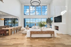 A spacious living room with a large dining table, comfortable seating, a fireplace, and large windows offering a view of the outdoors. - Lake Front Main House with Gulf Views, Pool and Low-Speed Vehicle (Santa Rosa Beach)