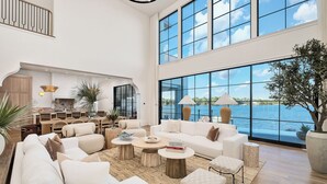 A spacious living room with a large window overlooking a body of water, a dining area with wooden furniture, and a staircase. - Newly Built | Pool | LSV | YOLO Boards (Santa Rosa Beach)