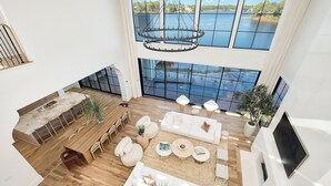 A spacious living area with a large window overlooking a body of water, a wooden dining table, and a modern chandelier. - Newly Built | Pool | LSV | YOLO Boards (Santa Rosa Beach)