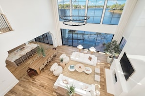 A spacious living area with a large window overlooking a body of water, a wooden dining table, and a modern chandelier. - Lake Front Main House with Gulf Views, Pool and Low-Speed Vehicle (Santa Rosa Beach)