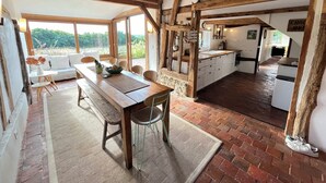Un interior rústico con una mesa de comedor de madera, una zona de cocina y ventanas amplias que dan a zonas verdes.