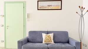 Et værelse med en lysegrøn dør, en grå sofa med pude, et indrammet landskabsmaleri og en høj vase med grene.