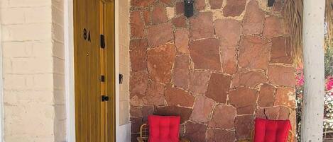 A covered porch with stone walls, a wooden door, and two red cushioned chairs.