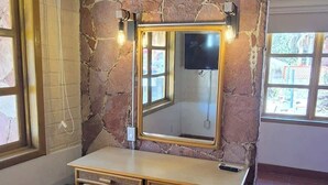 Ein Zimmer mit Steinwand, einem hölzernen Waschtisch mit Spiegel, einer Bank und einem Fenster.