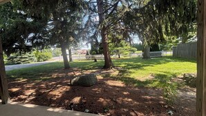 Un espacio al aire libre residencial con césped, árboles, una cerca blanca y una gran roca.