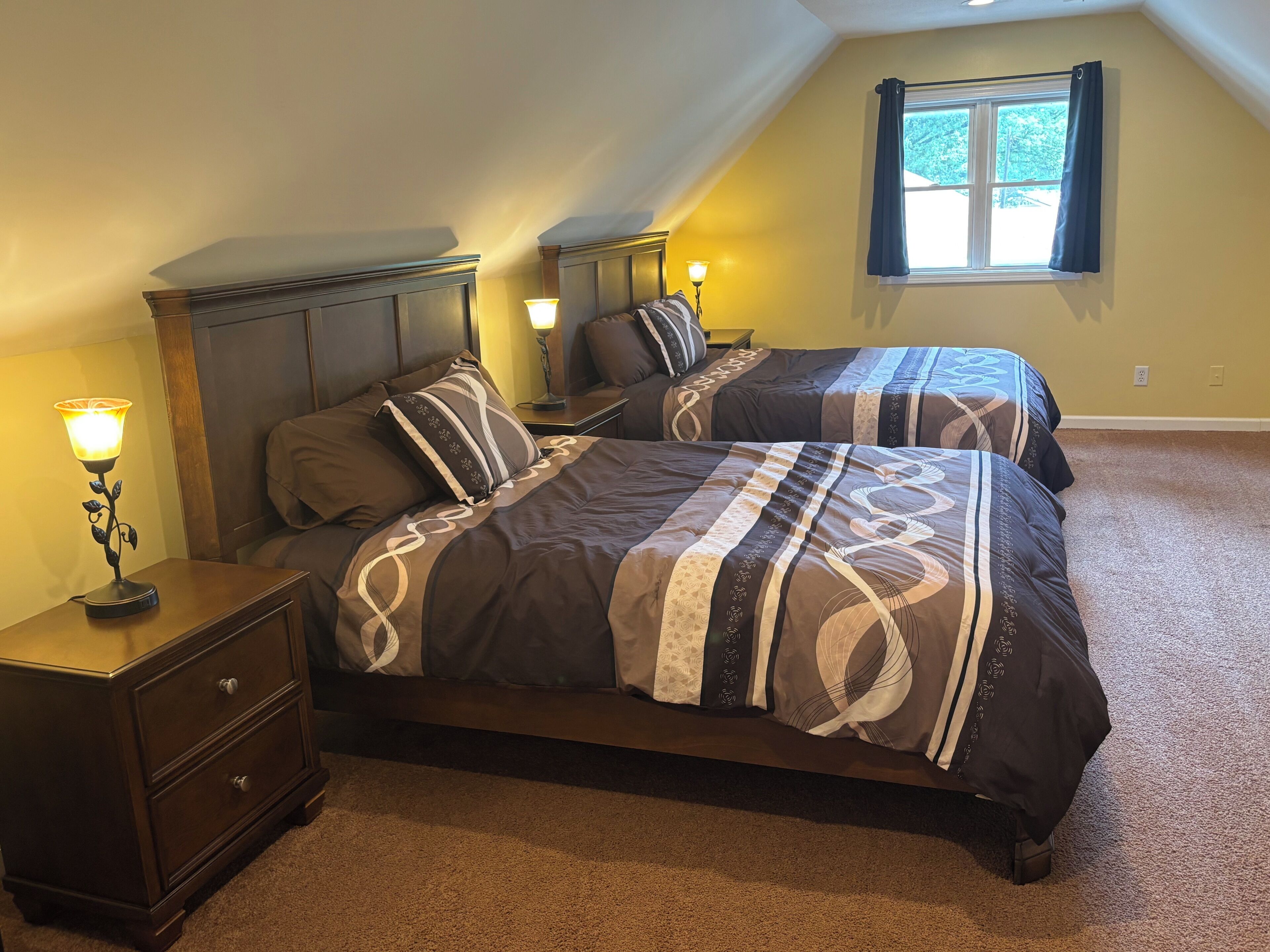 A bedroom with two beds, nightstands, a lamp, and a window.