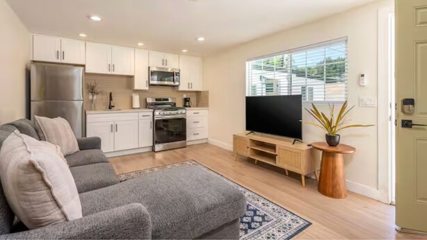 Eine moderne Küche mit weißen Schränken, einem Edelstahlkühlschrank und einer Mikrowelle. Ein graues Sofa, ein gemustertes Teppich und ein Flachbildfernseher auf einem Holzständer, sowie ein Topf mit Pflanze sind ebenfalls vorhanden.
