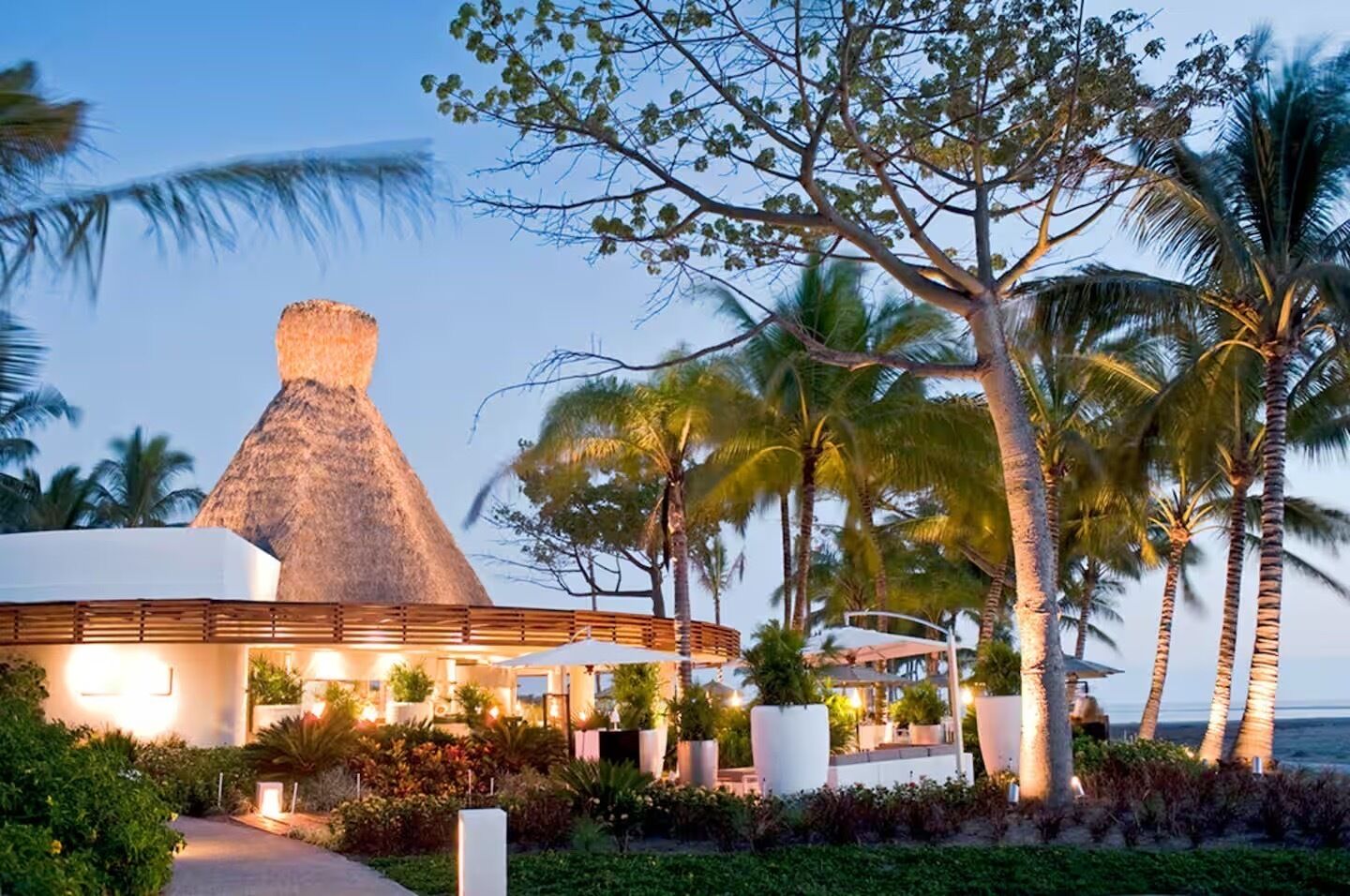 Ein Resort mit einem großen, mit Reet gedeckten Gebäude, Palmen und einem gut beleuchteten Außenbereich.