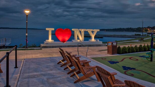 Blick auf das Wasserufer mit einem großen, beleuchteten 'I ♥ NY' Schild, Holzstühlen und einer Straßenlaterne.