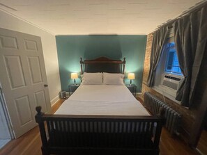 A bed with a wooden headboard, two bedside lamps, a window with curtains, and a radiator. - Uptown Geneva Rowhouse Flat (Geneva)