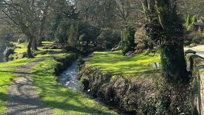 A serene pathway through a lush garden with a small stream, surrounded by trees and a stone wall. - Lakeview Villa (Liskeard)