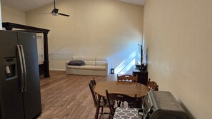 Eine kleine Küche mit Kühlschrank, einem Holztisch und Stühlen. Ein Sofa steht an der Wand, und ein Deckenventilator ist zu sehen.