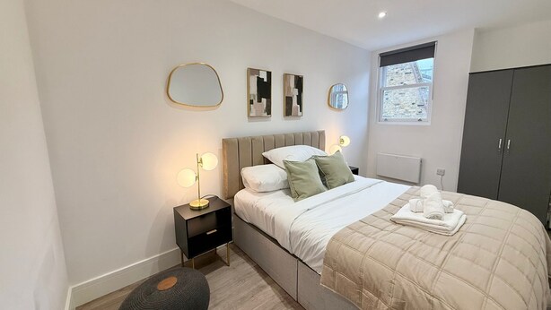 A modern bedroom with a bed, bedside tables, a nightstand, a mirror, and a wardrobe. - City Vibes & Comfort: 3BR Gem by Earl's Court (Greater London)