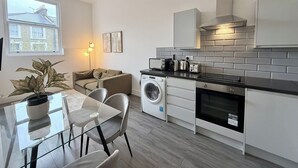 A modern kitchen with a washing machine, a dining area with a glass table and chairs, and a large window with a view of a house. - City Vibes & Comfort: 3BR Gem by Earl's Court (Greater London)