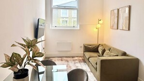 A small living room with a sofa, a dining table with chairs, a potted plant, a standing lamp, and a window with a view of a building. - City Vibes & Comfort: 3BR Gem by Earl's Court (Greater London)
