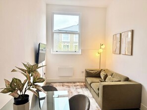 Ein kleiner Wohnraum mit einer Couch, einem Esstisch mit Stühlen, einer Topfpflanze, einer Stehlampe und einem Fenster mit Blick auf ein Gebäude.