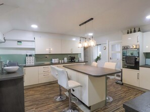 A modern kitchen with white cabinets, a central island with bar stools, a built-in oven, and pendant lighting. - Ty Crigyll (Holyhead)
