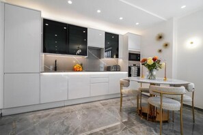 A modern kitchen with white cabinetry, a marble countertop, and a dining area with a round table and four chairs. - Cumberland Court (London)