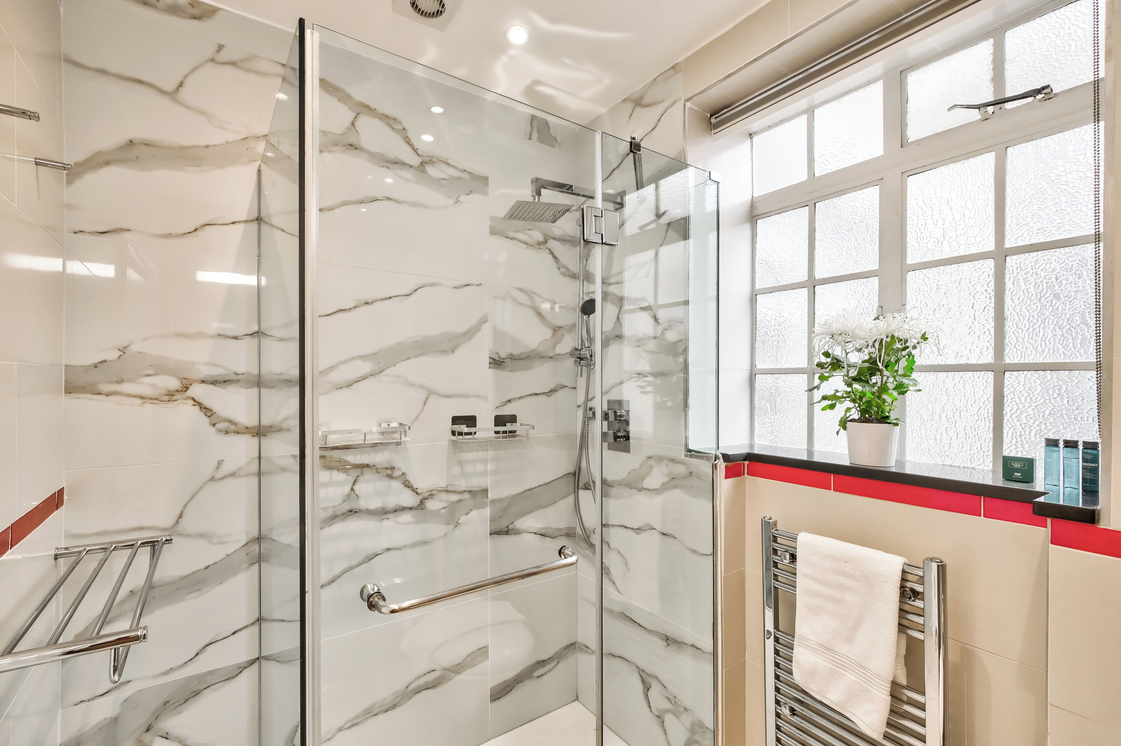 Un bagno moderno con pareti in marmo, una cabina doccia in vetro, una finestra con una pianta e un appendi-asciugamani.