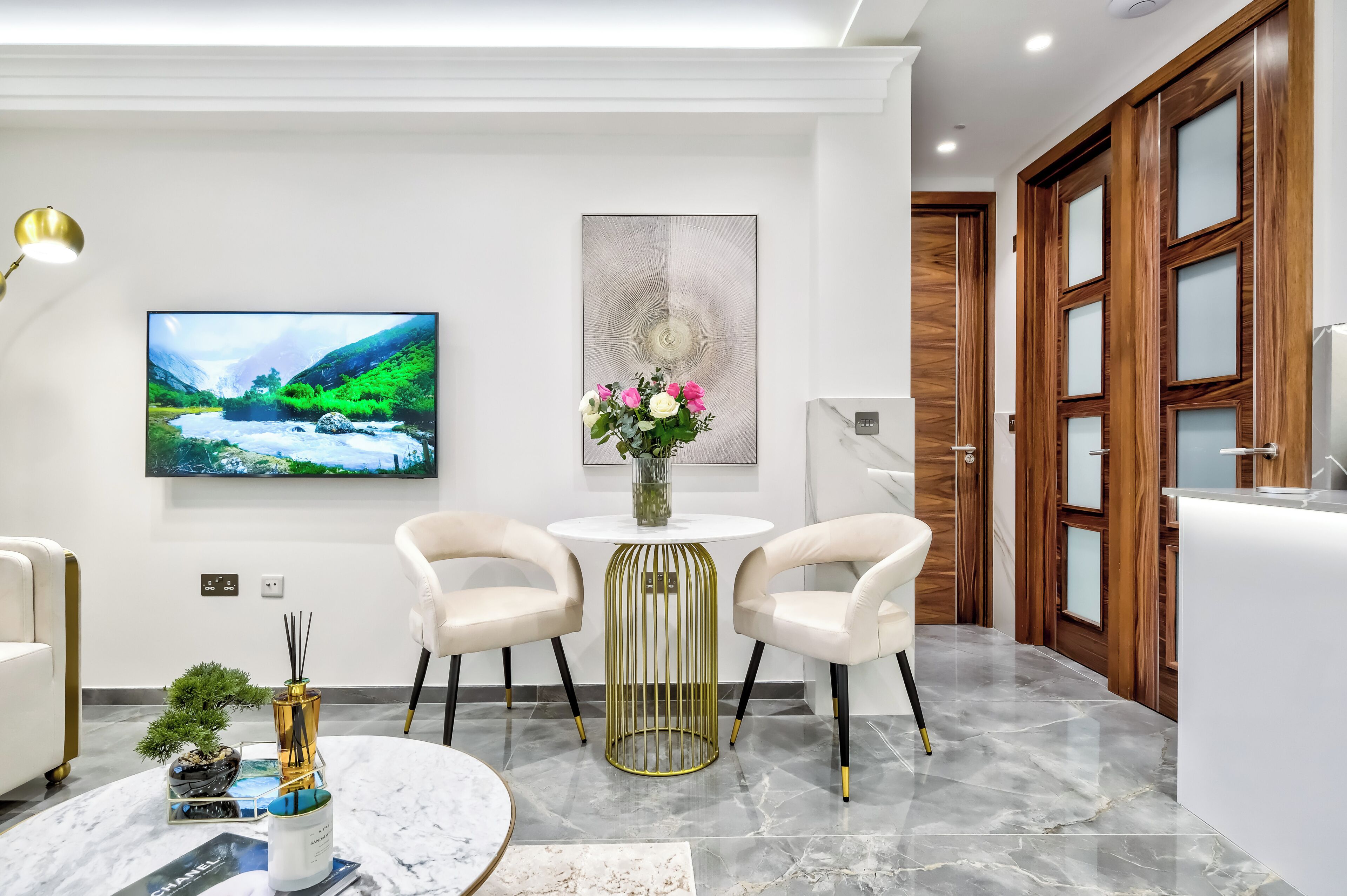 Una habitación de hotel moderna con un televisor de pantalla plana, una mesa redonda dorada con dos sillas, una mesita redonda con un florero y una puerta de madera grande.