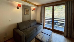 Ett vardagsrum med en soffa, ett soffbord i glas, två vägglampor, en tavla och en skjutdörr i glas som leder ut till ett utomhusområde.