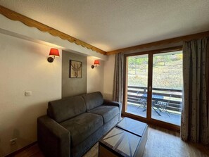 A living room with a sofa, a glass coffee table, two wall-mounted lamps, a painting, and a sliding glass door leading to an outdoor area. - Resort center, floor -1, fitness, swimming pool, sauna, balcony, tv, ski locker, 35m², Les Orres (Les Orres)