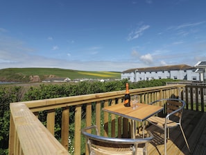 A wooden deck with a table and chairs, a bottle of champagne, and a glass. In the background, there is a building with a blue roof and a view of green fields and hills. - The Swift (St Bees)