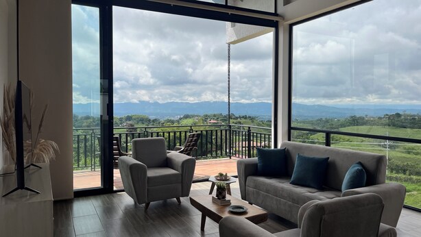 A modern living room with a sofa, armchairs, and a coffee table. Large windows offer a view of a balcony and mountains.